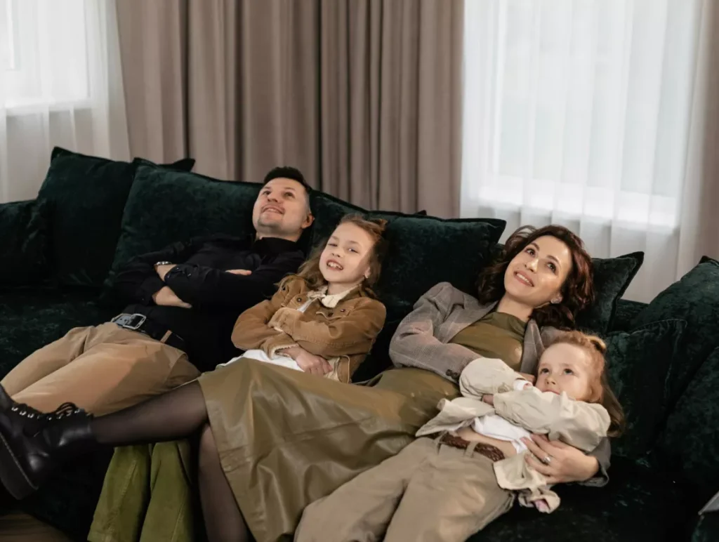 A family of four sits together on a couch, with a young child nestled between the parents, smiling and relaxed.